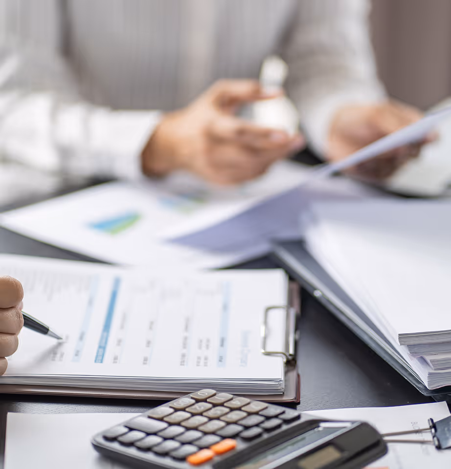 Close-up of a calculator and documents on a desk with a person writing on a clipboard in the background.