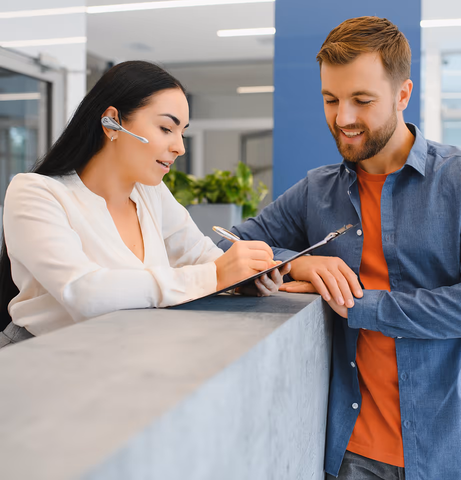 Receptionist wearing headset and white blouse helping a smiling man in a blue shirt and orange t-shirt by showing a clipboard.