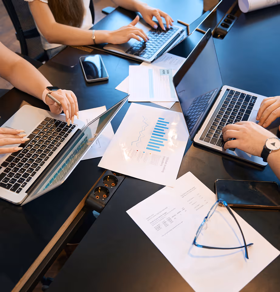 Three people working on laptops at a table with charts, papers, smartphones, and eyeglasses.