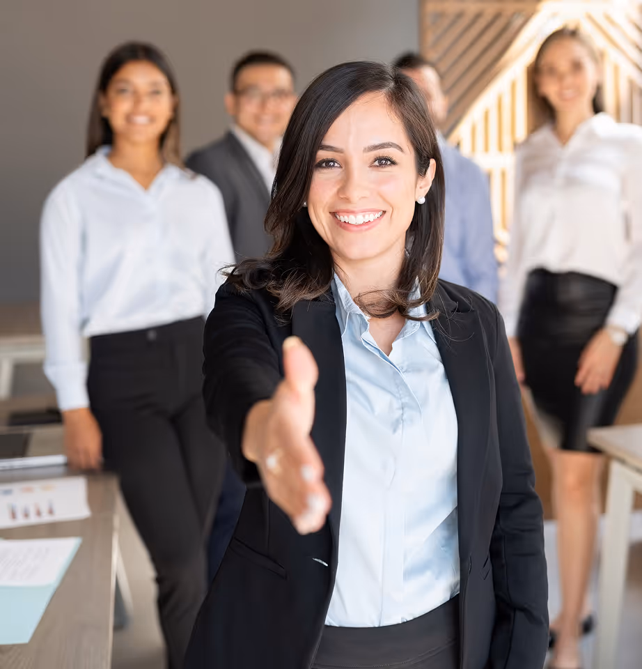 Businesswoman smiling and extending her hand for a handshake with three colleagues standing behind her.