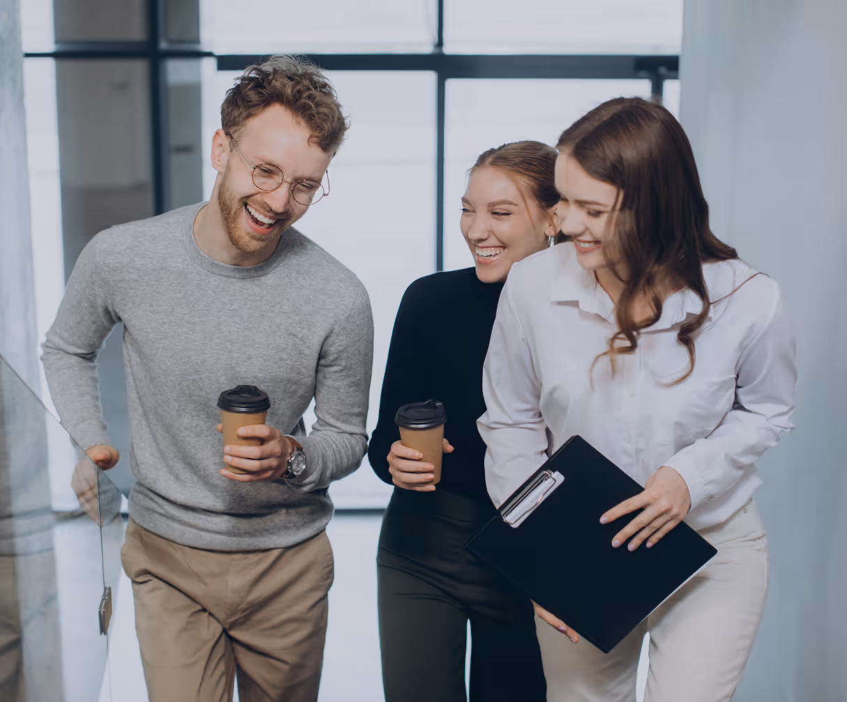 Three young professionals laughing and holding coffee cups and a clipboard while walking in an office hallway.