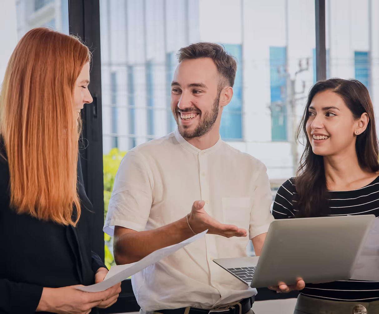 Three colleagues smiling and discussing work with a laptop and papers in an office.