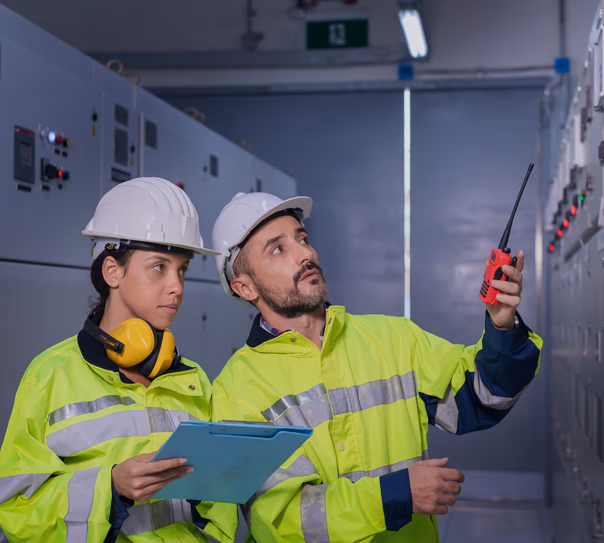 Two engineers in yellow high-visibility jackets and white helmets inspecting control panels, one holding a clipboard and the other a red walkie-talkie.