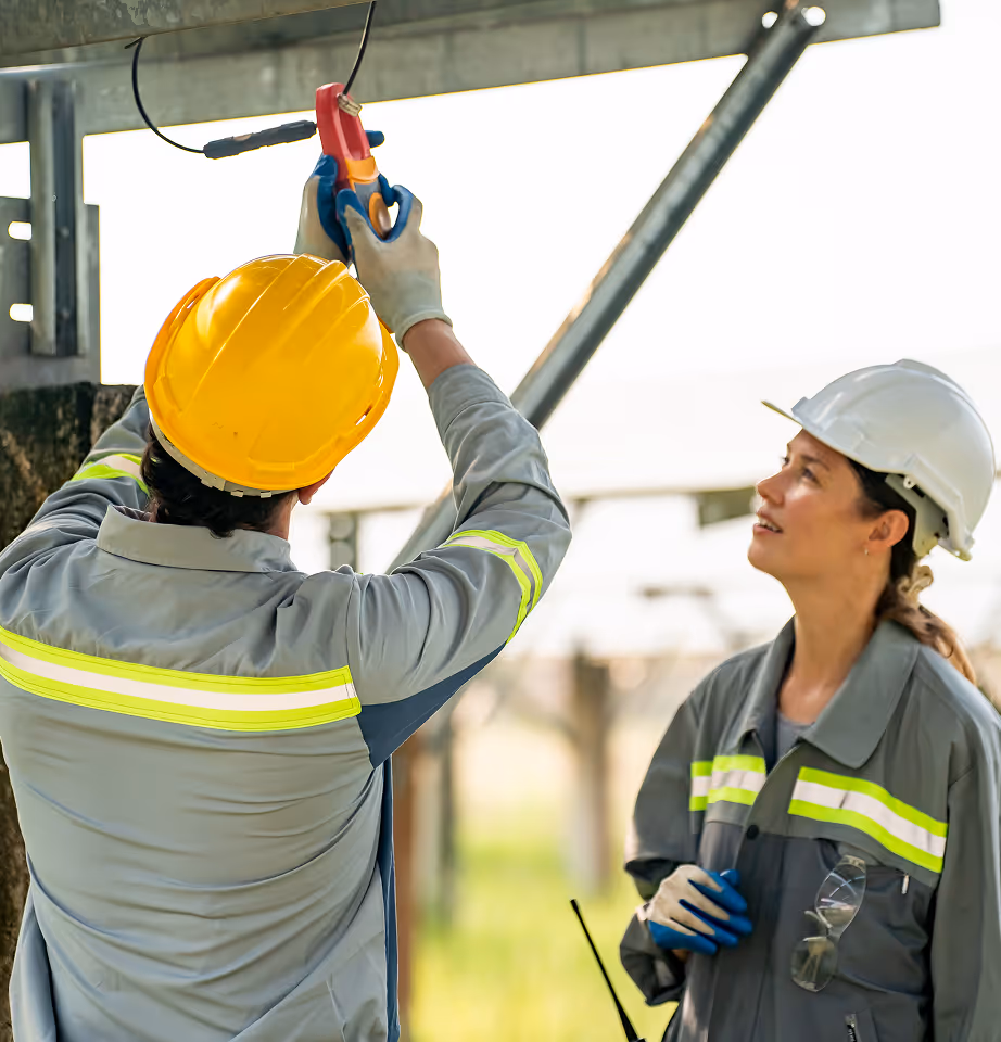 Two construction workers in safety helmets, one testing electrical wiring with a multimeter while the other observes.
