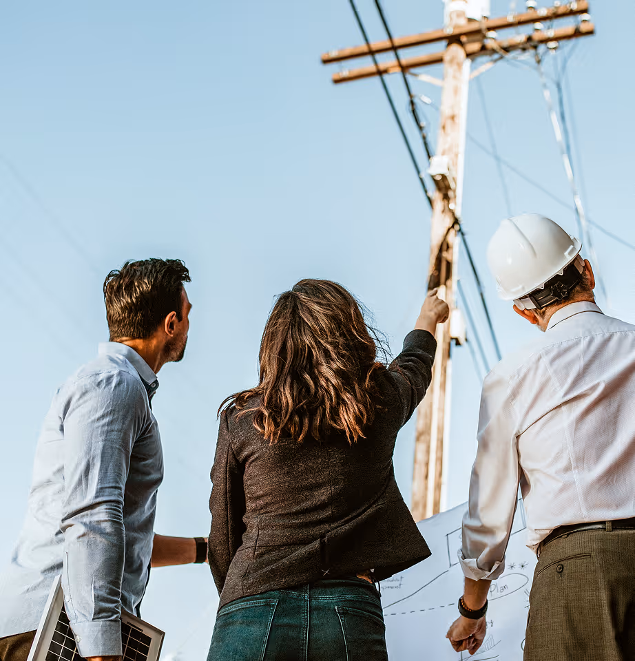Three professionals, including a woman pointing up, discuss plans in front of a utility pole with wires against a clear blue sky.