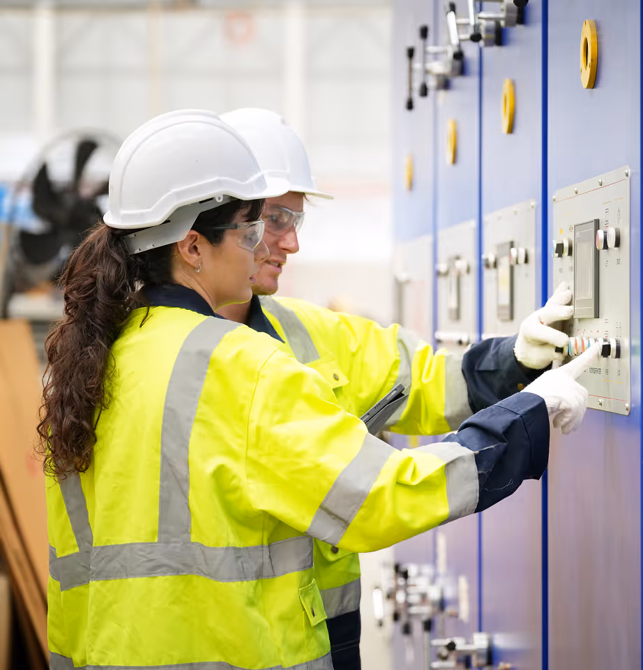 Two workers wearing yellow reflective jackets, hard hats, and safety glasses operating control panels on a blue industrial machine.