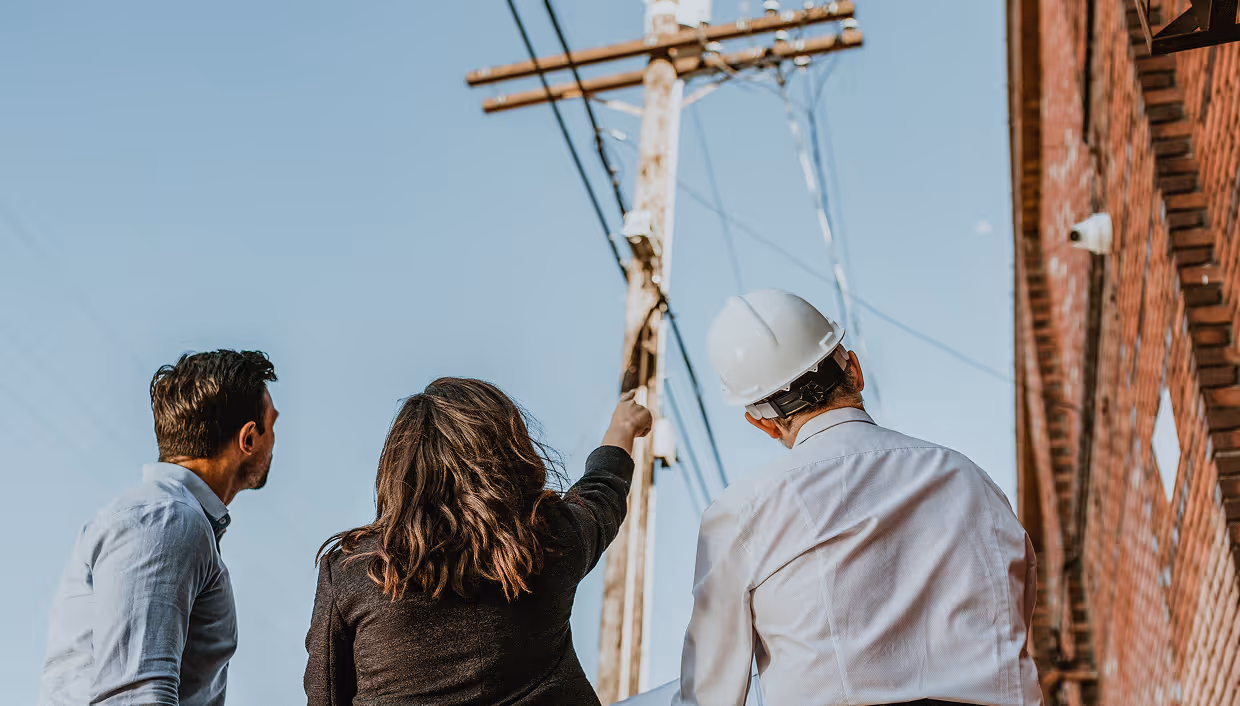 Three people, including a construction worker wearing a white hard hat, looking up and pointing at an electrical pole with wires against a clear sky.