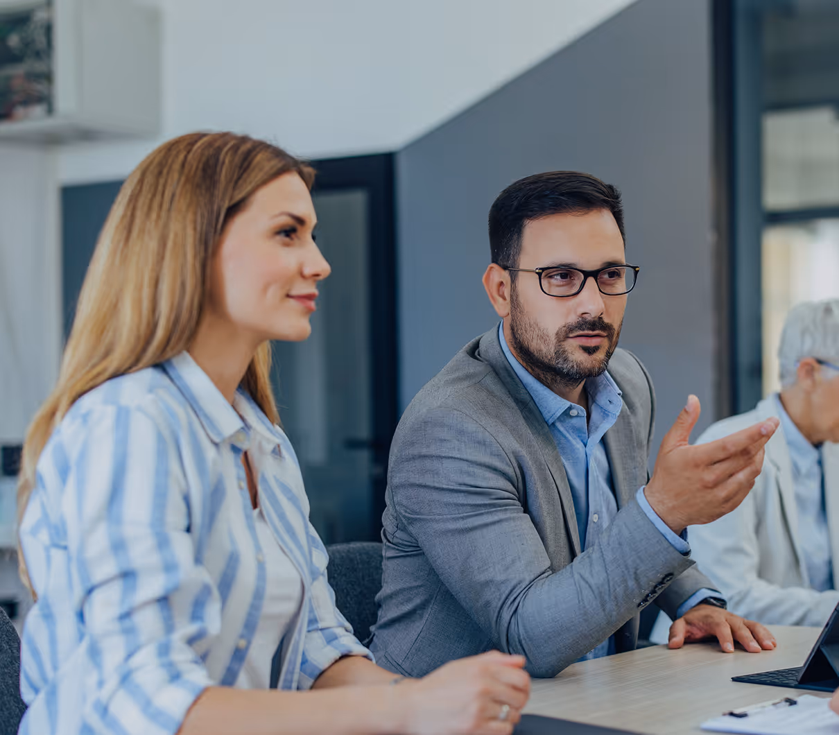A man in a gray blazer explaining something to a woman in a striped shirt during a meeting.