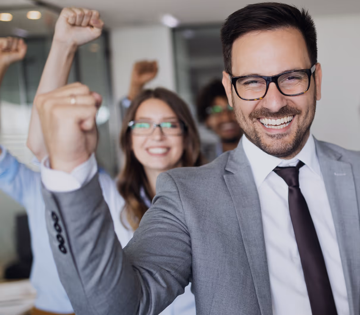 Smiling businessman in a gray suit and glasses raising a fist with blurred colleagues doing the same behind him.