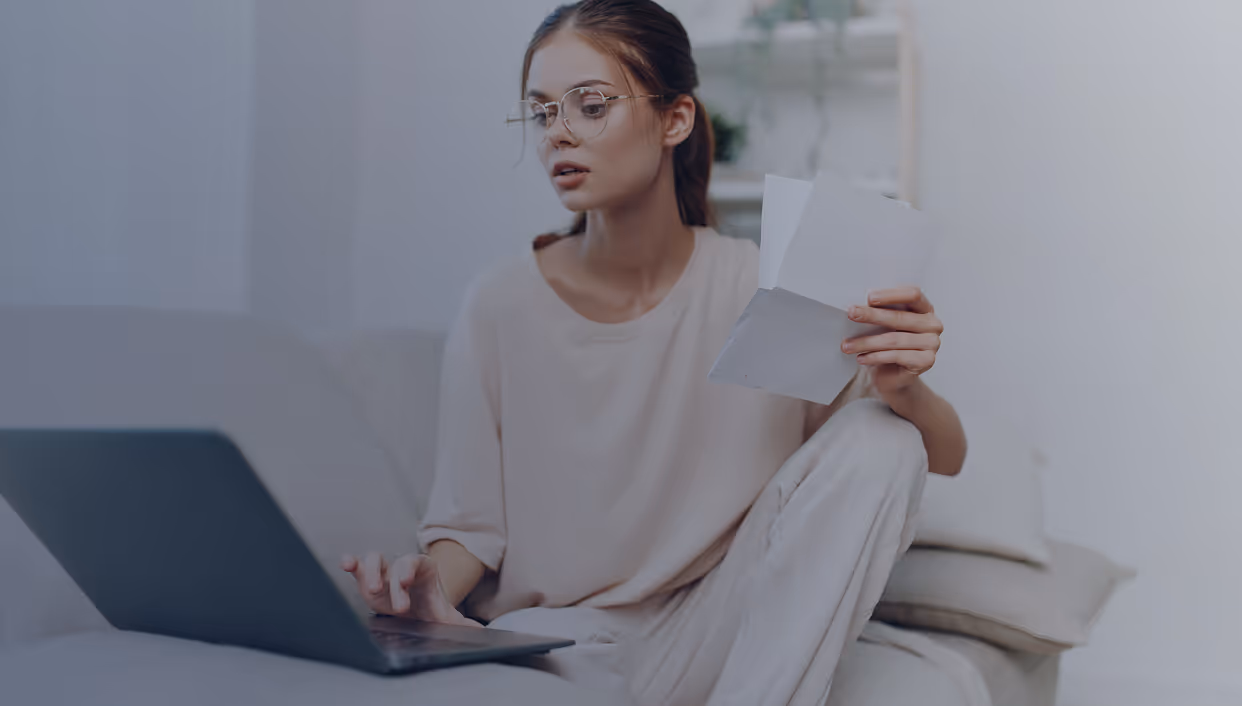 Young woman wearing glasses holding documents and using a laptop while sitting on a couch.
