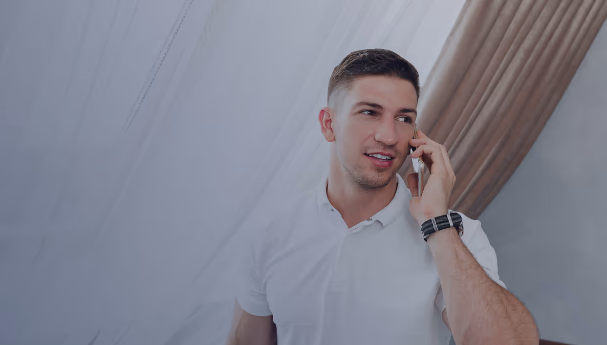 Man in white shirt talking on a smartphone with beige curtains in the background.