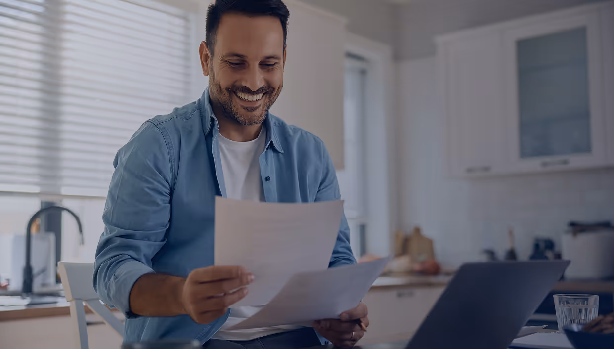 Smiling man sitting at a kitchen table reviewing papers with a laptop nearby.