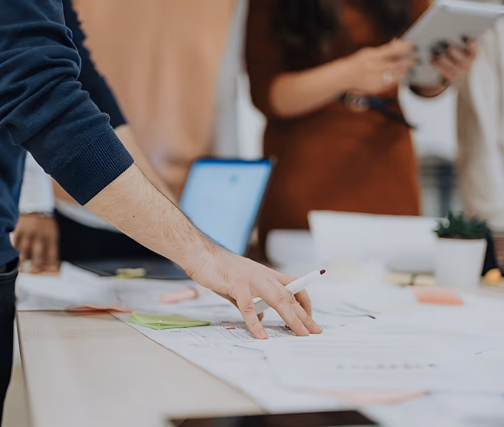 Person's hand holding a marker pen over a table with architectural plans, with blurred people and laptop in the background.