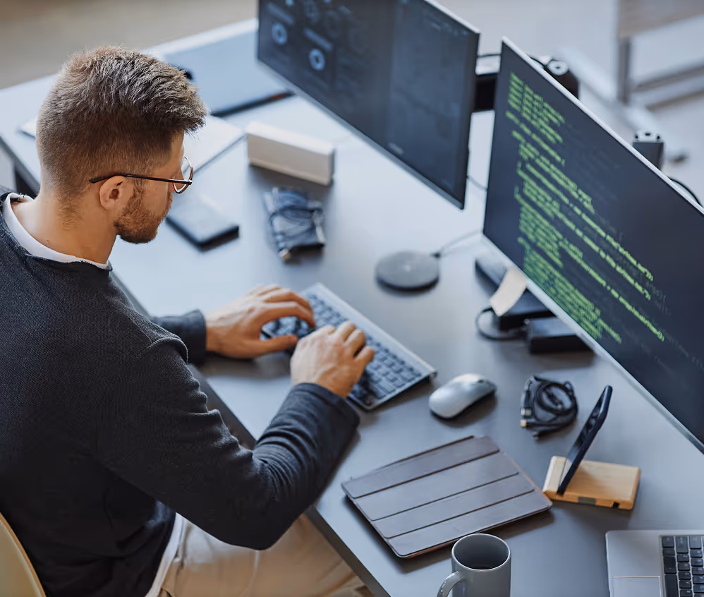 Man with glasses typing on a keyboard at a desk with dual monitors displaying code.
