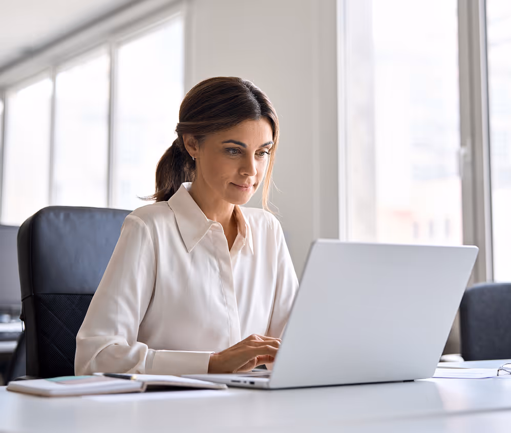 Woman in a white blouse working on a laptop at an office desk with notebooks and a pen.