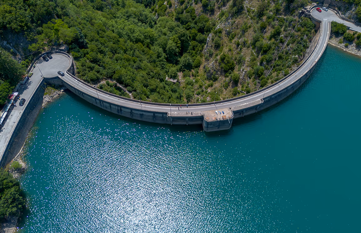 Aerial view of a curved dam with a road on top, surrounded by green wooded hills and turquoise water reflecting sunlight.