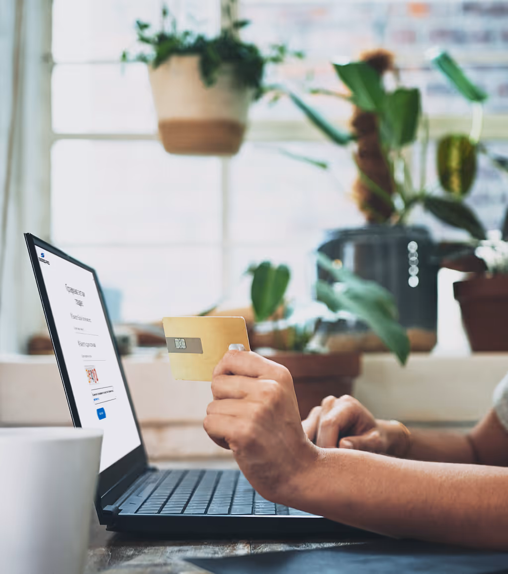 Ръка държи златна кредитна карта пред лаптоп с отворен екран за онлайн плащане, на заден план са стайни растения.