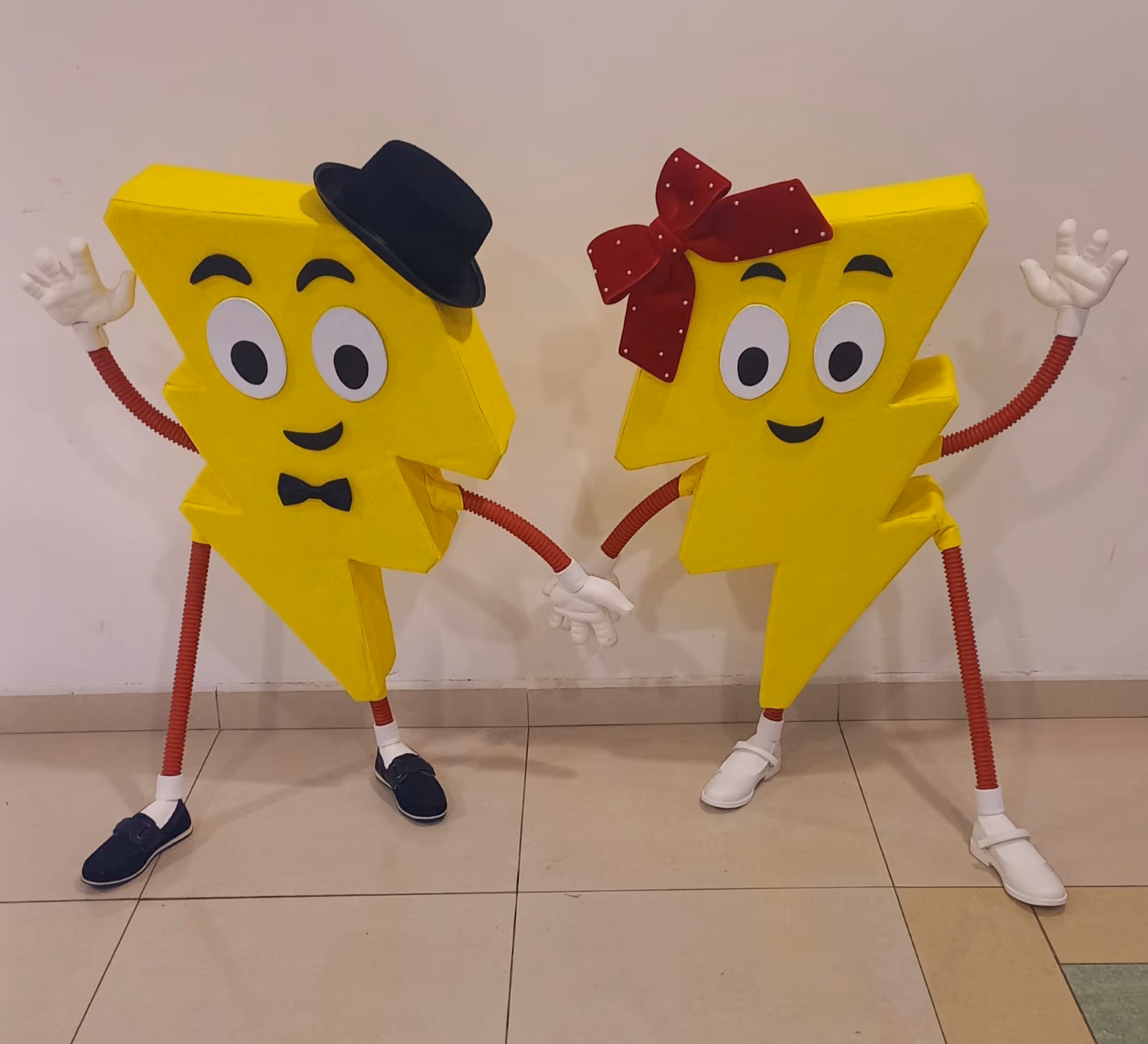 Две жълти костюми на светкавици с усмихнати лица, черна шапка и папионка на едната, червен панделка на другата, държащи се за ръце.