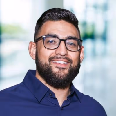 Smiling man with glasses and a beard wearing a dark blue collared shirt, with a blurred indoor background.