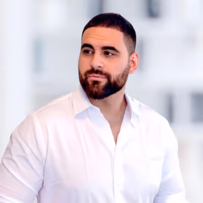 Man with a beard and short hair wearing a white button-up shirt, looking to the side against a blurred light background.