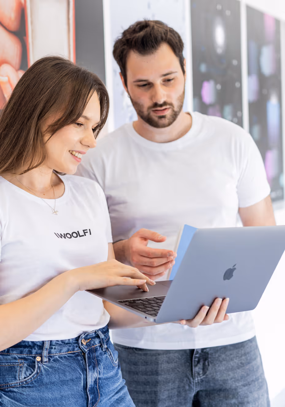 A candid photograph of four young adults collaborating around a MacBook laptop on a tabletop in a modern office or study space. A smiling woman in the foreground, wearing a white t-shirt with the word "WOOLF" printed on it, operates the laptop. Another woman looks at the screen from the right, and two individuals (a man with dark, curly hair and mustache, and a third person) stand behind them, observing the screen. The background is brightly lit with large windows or glass partitions.
