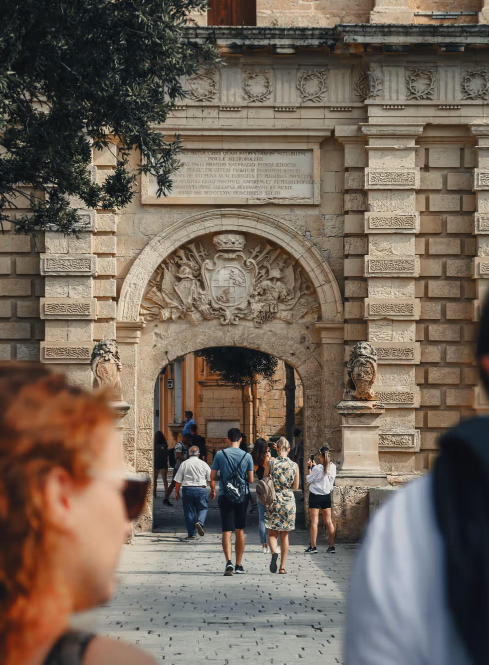 A slightly out-of-focus, street-level photograph looking through a large, ancient, stone arched gateway in an old European city. 