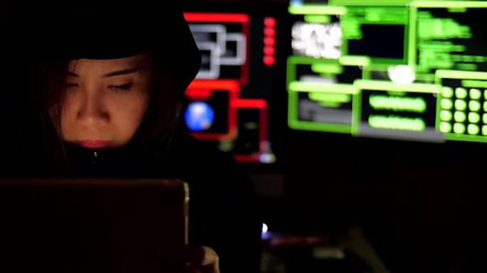 A person in a hoodie looks at a device in a dim room, with brightly lit computer screens displaying green and red code in the background.