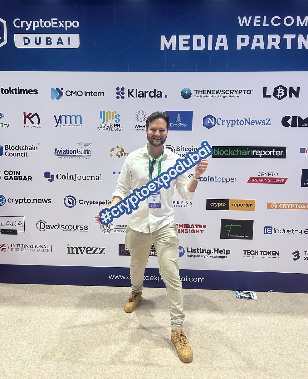 Dennis smiling at CryptoExpo Dubai holding "#cryptoexpodubai" sign. Background has a media partner board with various logos.