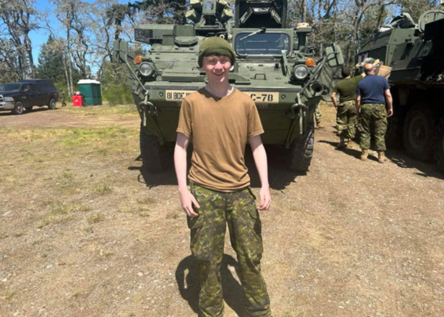 Young person smiling in military gear stands in front of an armored vehicle outdoors. Others in similar gear nearby. Sunny day, trees in background.