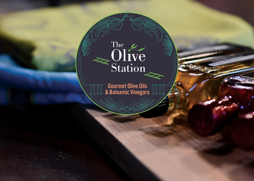 Bottles of olive oil and balsamic vinegar on a wooden board with The Olive Station logo overlay.
