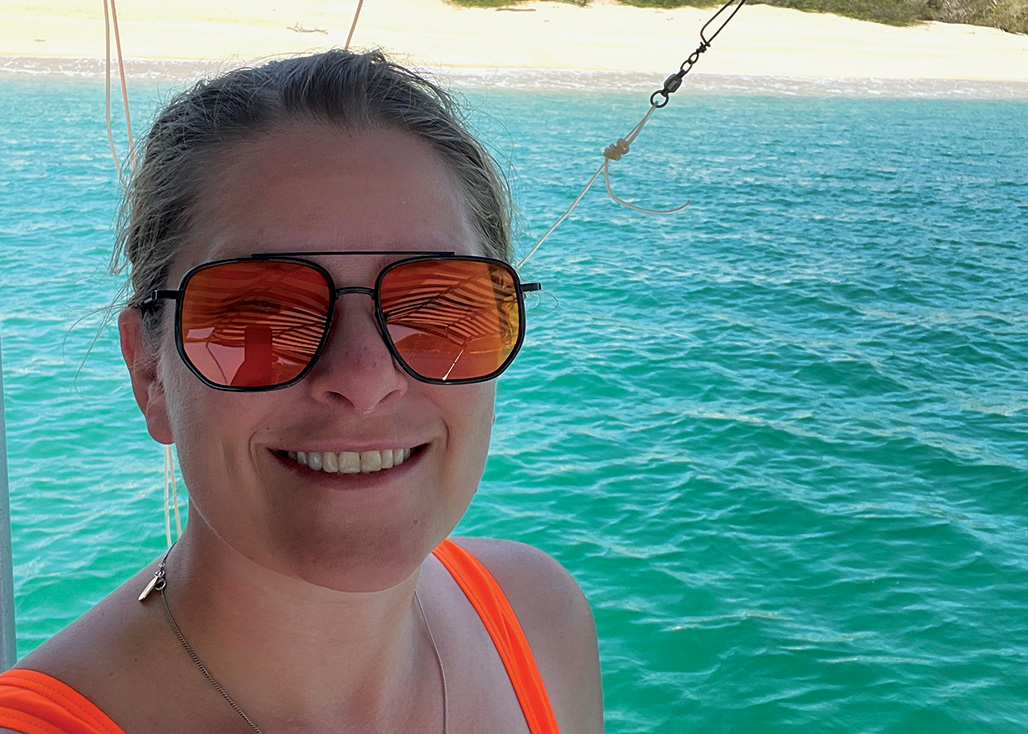 Smiling woman wearing orange sunglasses and top with ocean and sandy shore in the background.