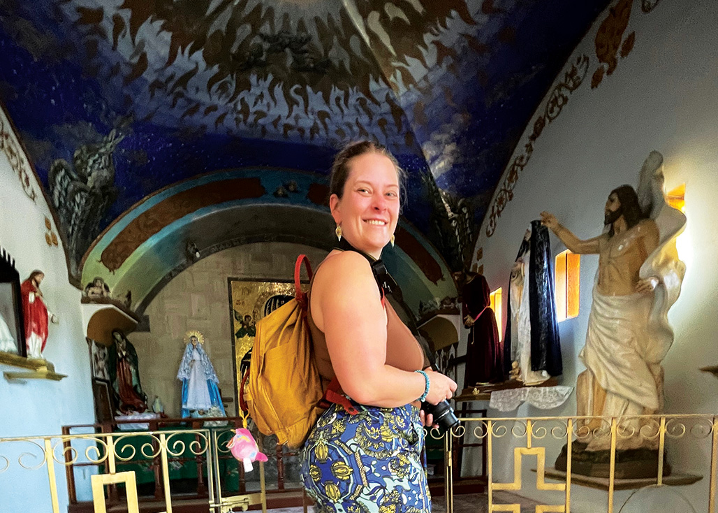Smiling woman with yellow backpack holding a camera inside a chapel with religious statues.