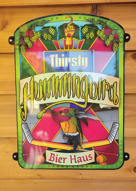 Colorful sign reading Thirsty Hummingbird Bier Haus with a hummingbird and hockey sticks on wood wall.