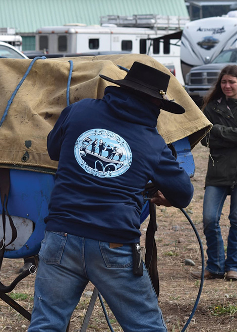 Man wearing black hat and hoodie with logo working on a horse packing demo. A woman watches nearby.