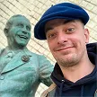 Un homme portant une casquette bleue pose devant une statue en bronze souriante.