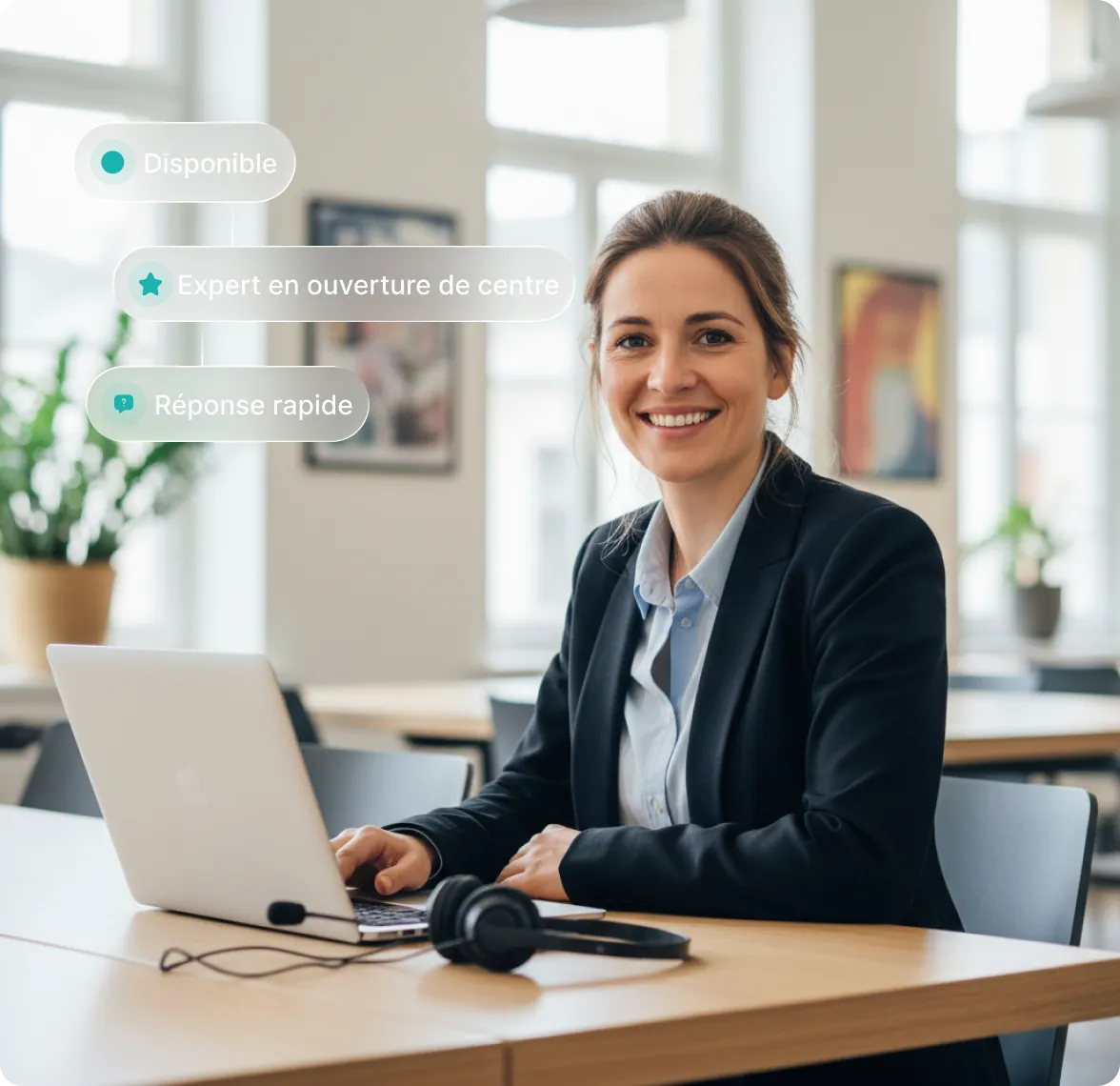 Femme souriante en costume assise à une table avec un ordinateur portable et un casque dans un bureau lumineux.