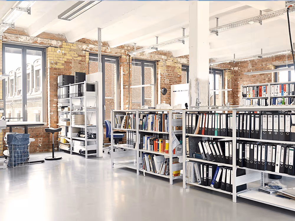 Bild Helles Büro mit weißen Regalen voller Ordner und Bücher, großen Fenstern und freiliegenden Ziegelwänden.