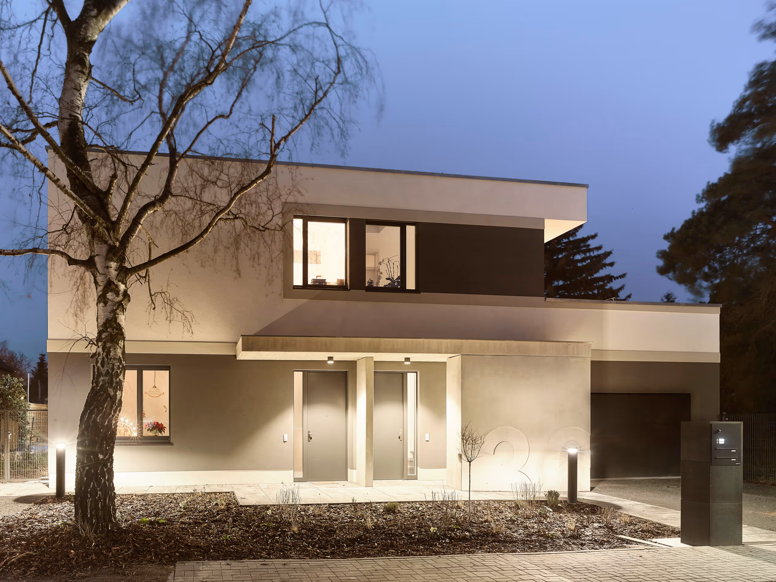 Modernes zweistöckiges Haus mit beleuchteten Fenstern, zwei Eingangstüren und einem Baum im Vordergrund bei Dämmerung.