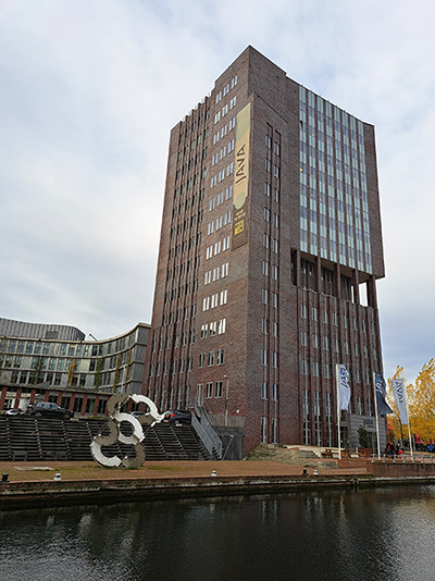 Hoog kantoorgebouw van baksteen met grote ramen aan het water en een metalen kunstwerk op de voorgrond.