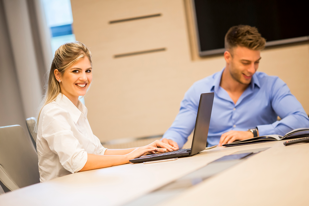 Twee jonge professionals, een vrouw in een witte blouse die lacht naar de camera en een man in een blauwe blouse die glimlacht terwijl hij naar een open boek kijkt, zittend aan een bureau met een laptop.