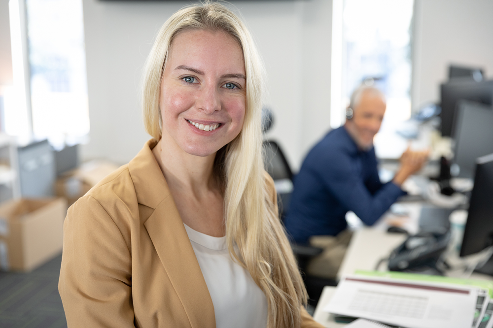 Blonde vrouw met beige colbert glimlacht in een modern kantoor met een man aan een bureau op de achtergrond.