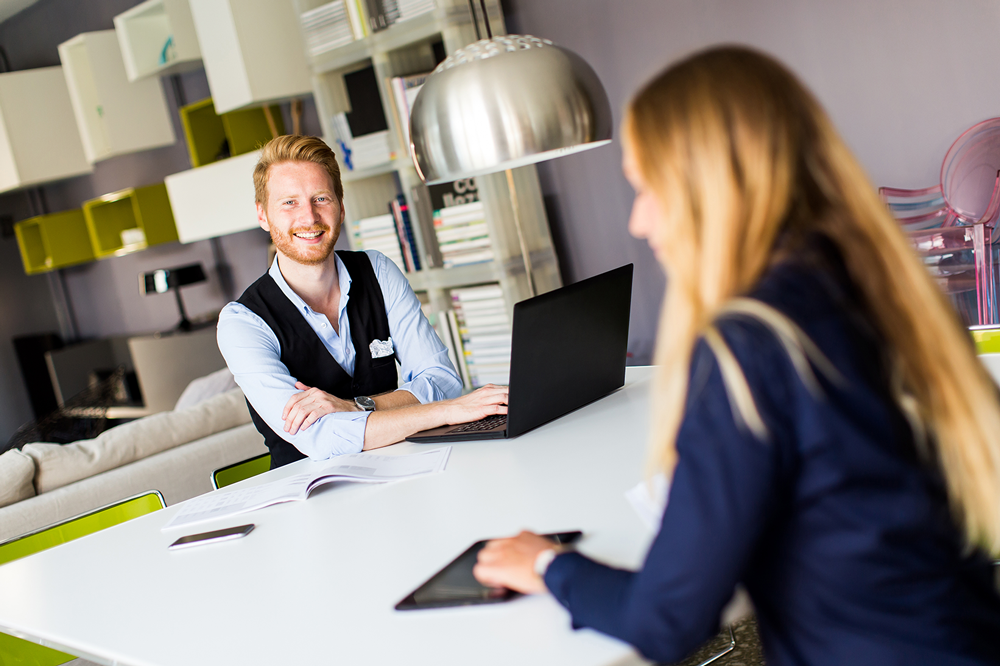 Een glimlachende man met een zwart vest werkt op een laptop aan een witte tafel tegenover een vrouw met lang blond haar in een moderne kantoorruimte.
