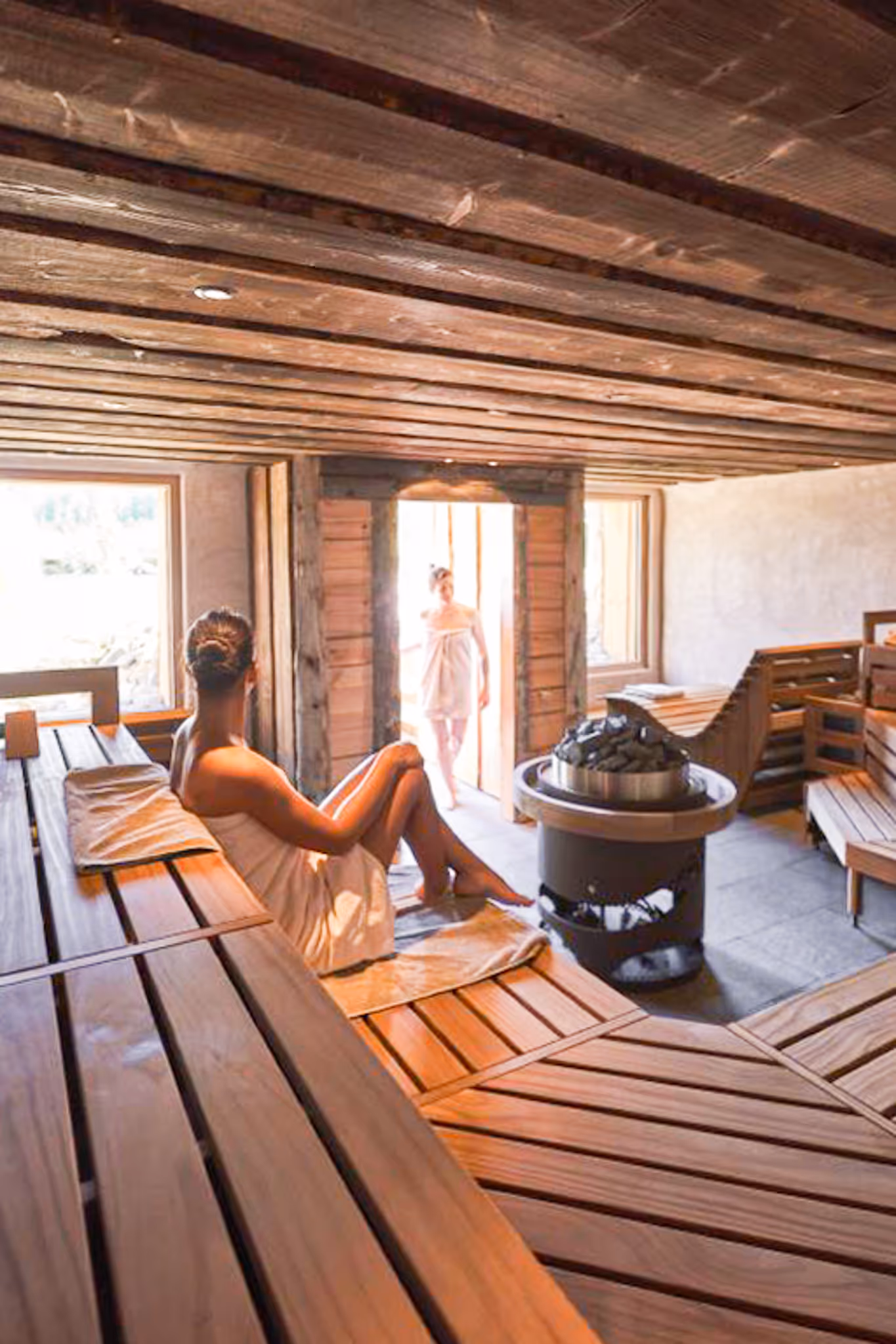Innenansicht einer Sauna mit zwei Frauen, die in Handtüchern auf Holzliegen entspannen, eine am Eingang stehend und eine sitzend.