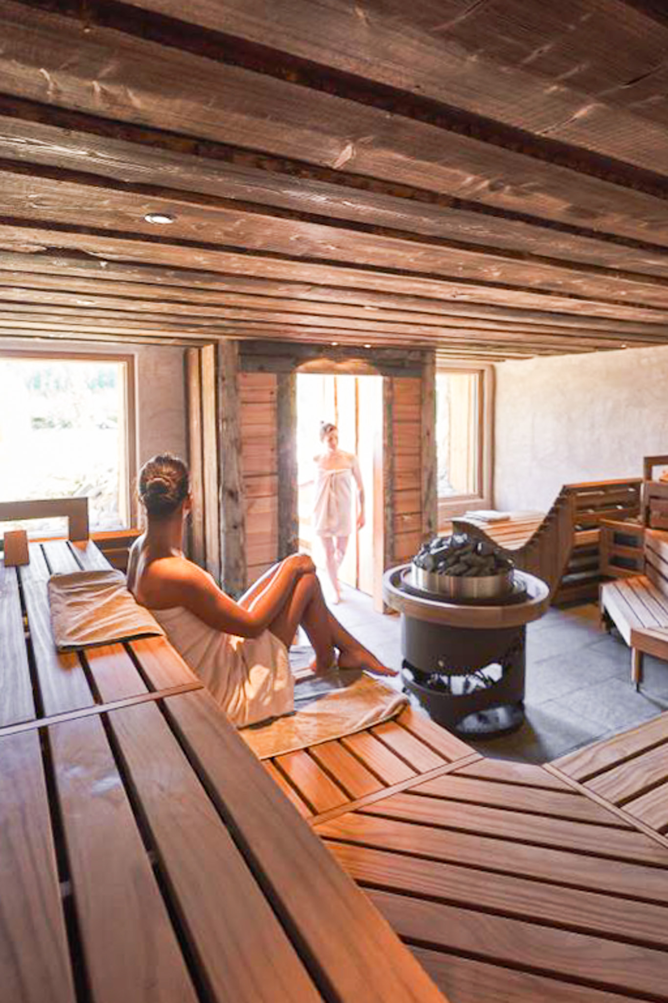 Innenansicht einer Sauna mit zwei Frauen, die in Handtüchern auf Holzliegen entspannen, eine am Eingang stehend und eine sitzend.