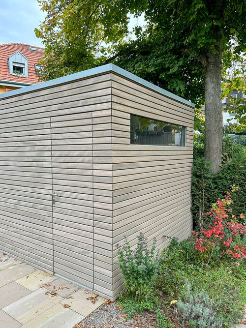 Moderner Gartenhaus aus horizontalen Holzlatten mit einem schmalen Fenster, umgeben von Bäumen und Gartenpflanzen.