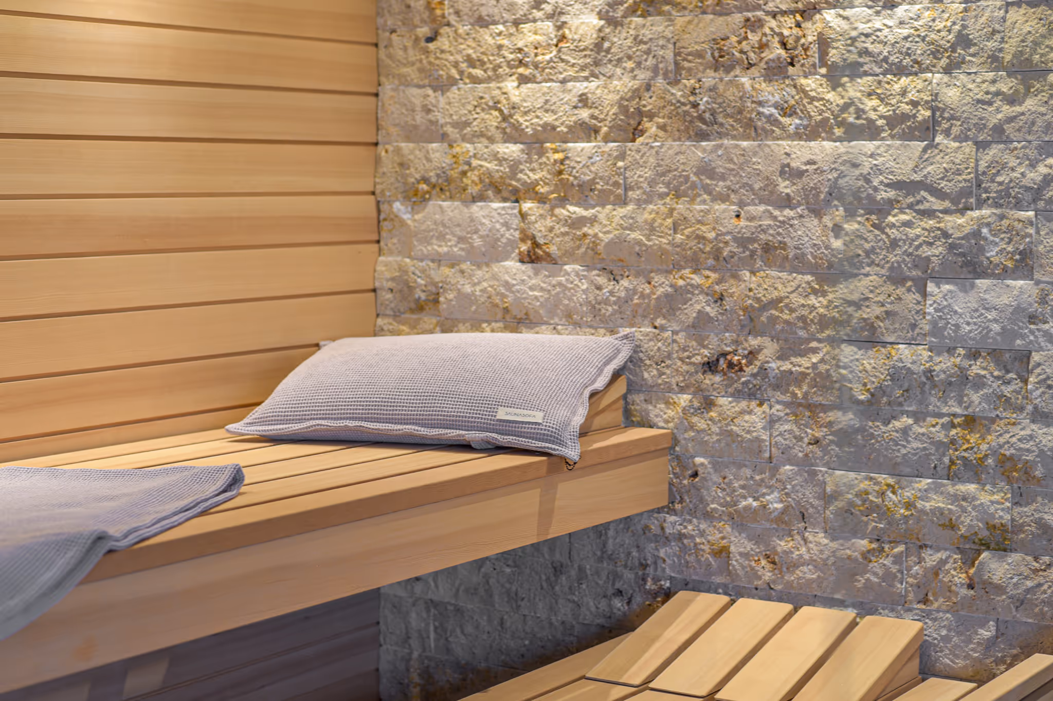 Holzbank in einer Sauna mit beigem Kissen, hellen Holzwänden und einer Steinwand im Hintergrund.