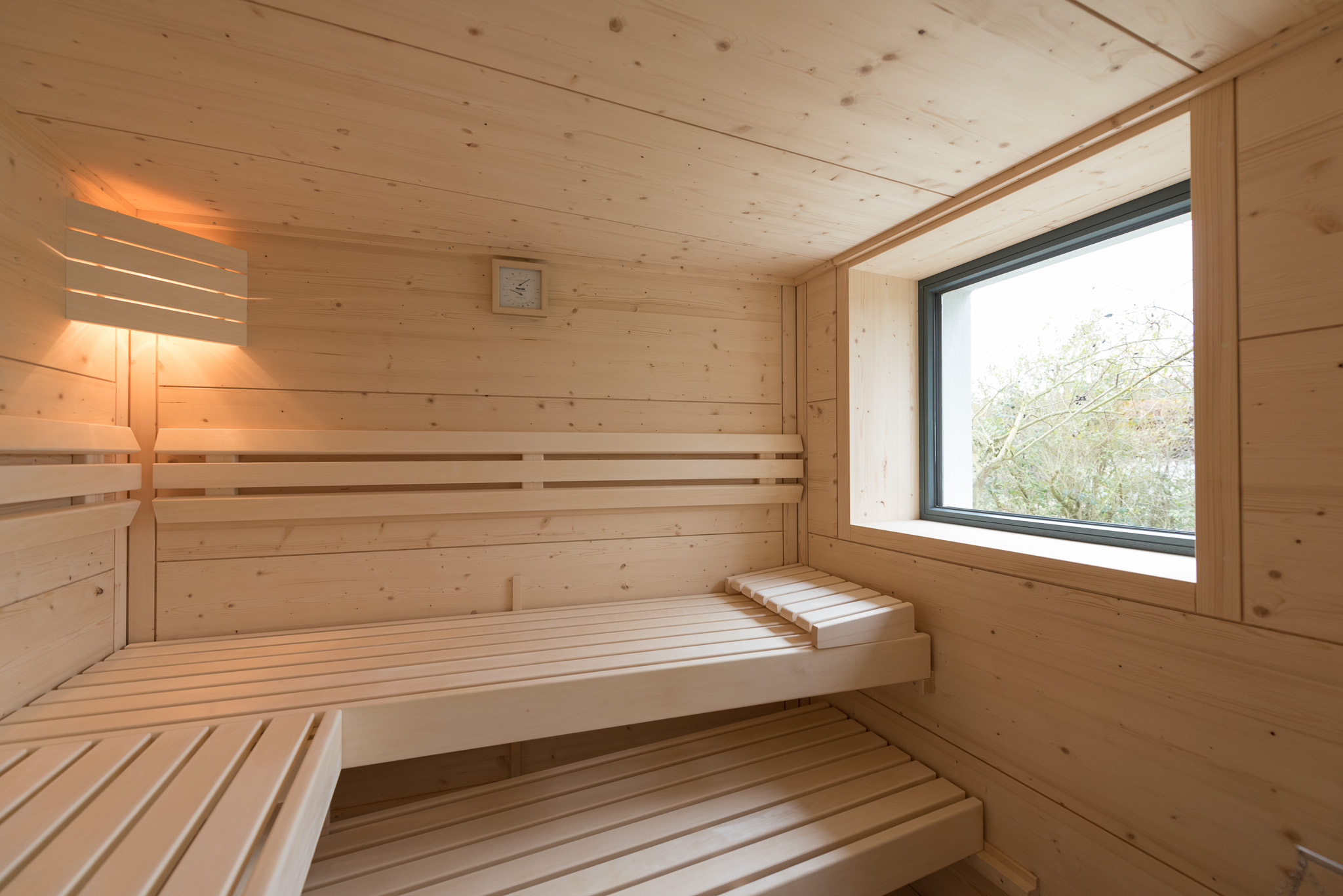 Innenraum einer Holzsauna mit zwei Etagen angrenzender Bänke, einem Fenster mit Blick auf grüne Bäume und einer beleuchteten Wandlampe.