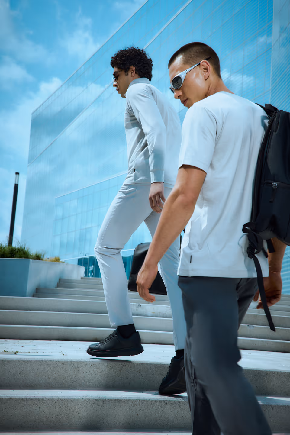 Two men wearing sunglasses and casual clothing walking up outdoor steps near a modern glass building under a blue sky.