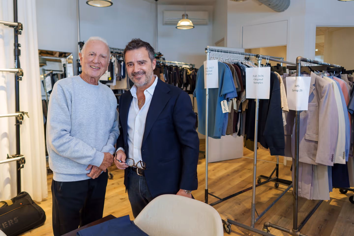 Two men smiling and standing together in a clothing showroom with racks of garments behind them.