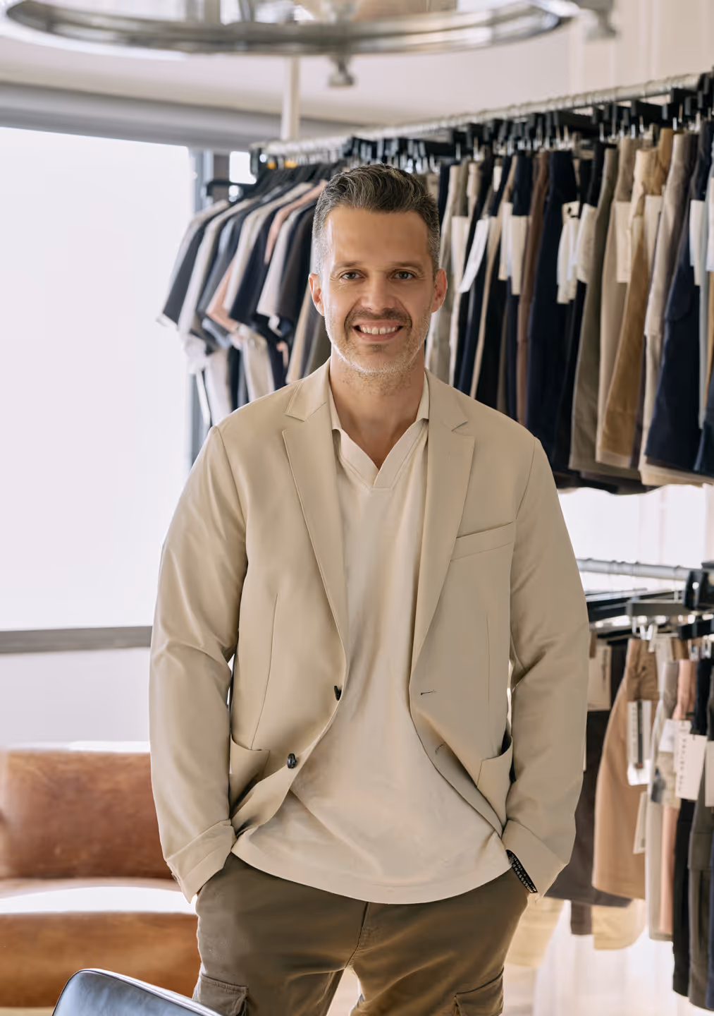 Smiling man in beige blazer and shirt standing in a clothing store with racks of pants and shirts behind him.